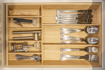 Organized kitchen drawer with stainless steel cutlery and utensils in a wooden divider. Neatly arranged spoons, forks, knives, and kitchen tools such as a corkscrew, garlic press, and bottle opener 