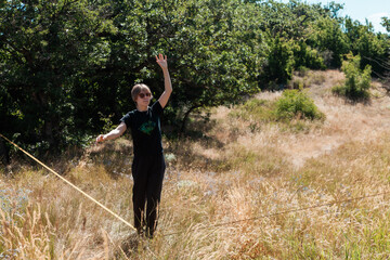 A female slackliner performs acrobatic poses and splits on a slackline stretched between trees on a mountain hilltop in Georgia. Aerial views, dry summer grass, and scenic peaks surround her