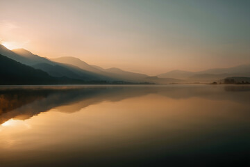 Fototapeta premium Minimalist sunrise landscape with misty lake and soft mountain layers, perfect as background or with copy space for text and design