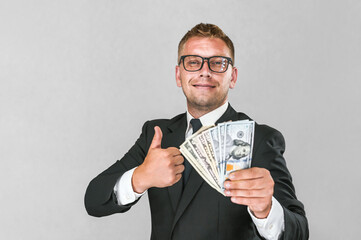 Cheerful businessman in a suit holds american dollars in his hands and thumbs up on a gray and...