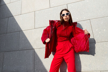 High fashion photo of a young brunette woman in red glossy down jacket, and red pants. Urban street...