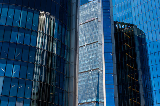 Reflection of neighboring towers on blue-tinted glass facade, showing transparency, geometry and light in a dense vertical business cluster.