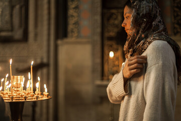 Woman with Asian-European features in headscarf and white dress praying and lighting candles in an...