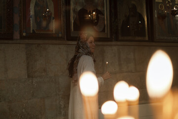 Woman with Asian-European features in headscarf and white dress praying and lighting candles in an...