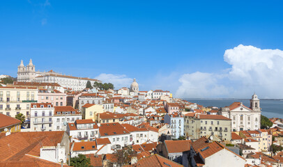Colorful Lisbon cityscape skyline with red rooftops cascading down to the Tagus River under a bright blue sky with fluffy white clouds.