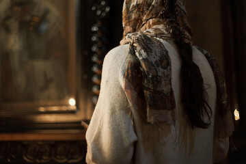Woman with Asian-European features in headscarf and white dress praying and lighting candles in an Orthodox church. A quiet, spiritual moment of faith and tradition