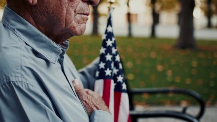 Elderly veteran stands respectfully with his hand over his heart, near a waving american flag in a serene park setting, honoring his service and the nation - Powered by Adobe