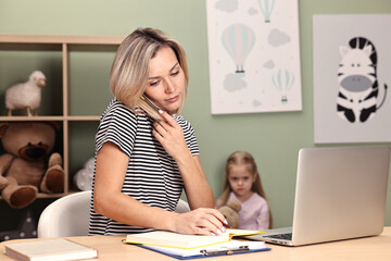 Work-family balance. Single mother talking by smartphone at table and her offended daughter in...