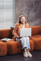 Fototapeta premium Smiling entrepreneur works at home on a laptop while comfortably seated on a stylish sofa in a tranquil indoor space. Working from home, woman in business jacket and pajama pants and slippers