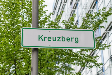 Stra&szlig;enschild mit der Aufschrift &bdquo;Kreuzberg&ldquo; in gr&uuml;ner Schrift, vor einem Hintergrund aus gr&uuml;nen B&auml;umen und moderner Glasarchitektur in Berlin