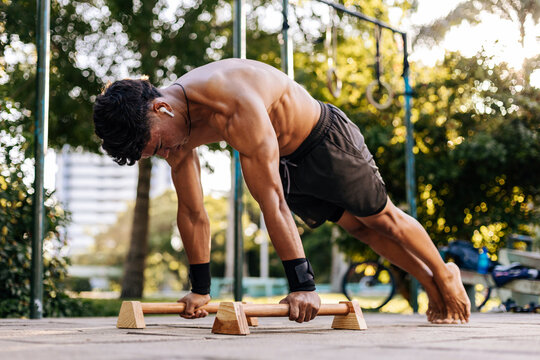 Young man doing calisthenics push-ups on wooden parallettes at an outdoor park. Shirtless athlete trains upper body strength with focus and control. Urban fitness and bodyweight workout