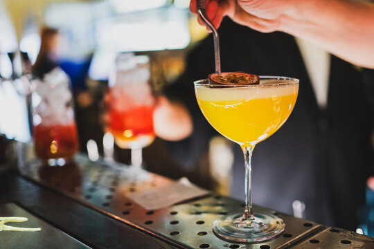 A bartender garnishes a vibrant yellow cocktail with a halved passion fruit in a coupe glass, set on a stylish bar counter, creating a refreshing and exotic drink experience.