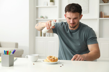 Emotional young man spraying insecticide onto pretzel with cockroaches at white table indoors. Pest problem