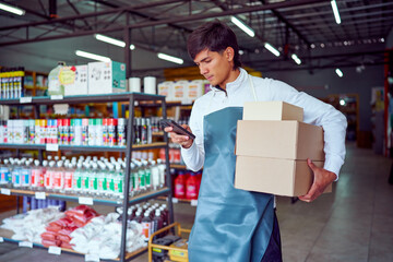 Latin american shopkeeper managing inventory using a smartphone while carrying boxes in a hardware store