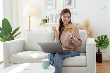 Young Woman Joyfully Shopping Online From a Cozy Living Room With a Laptop and Credit Card in Hand