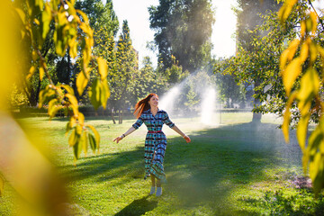 A happy young woman is joyfully walking through a sunny park where sprinklers are actively watering the grass