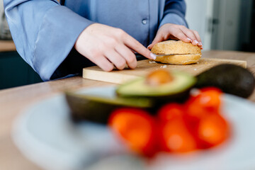 Teen girl's hands slicing bread roll near avocado and tomatoes in cozy kitchen
