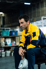 Warehouse worker wearing yellow uniform sitting on chair holding safety helmet appearing tired...