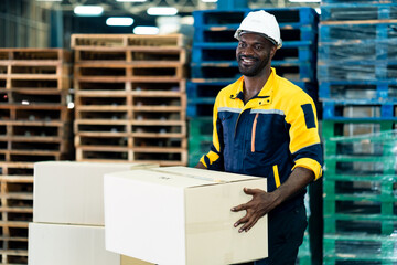Smiling African warehouse worker holding cardboard box wearing safety jacket and helmet standing in shipping area, adult male logistics staff lifting package, teamwork and delivery service concept