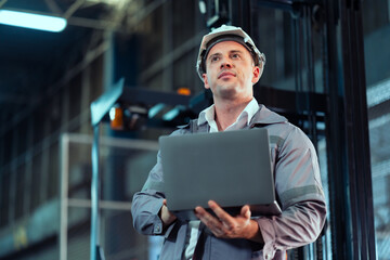 Male warehouse supervisor wearing gray jacket and helmet holding laptop inside industrial storage area, adult Caucasian man monitoring logistics operation workflow and facility efficiency