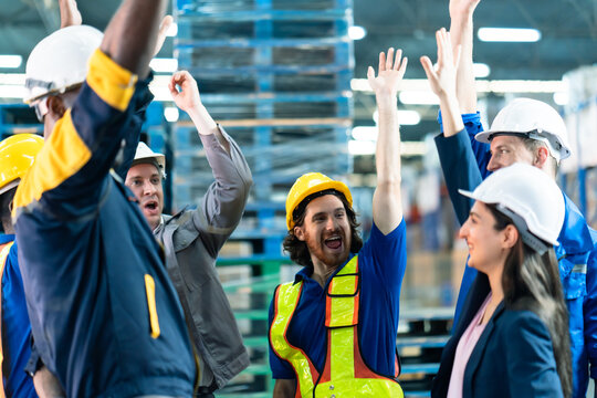 Logistics team raising hands in celebration inside warehouse, male and female workers in helmets and safety uniforms expressing excitement and motivation, industrial workplace unity and teamwork theme