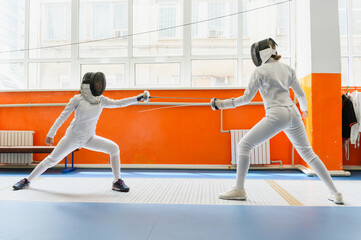 Female fencers lunging toward each other in bright fencing hall.