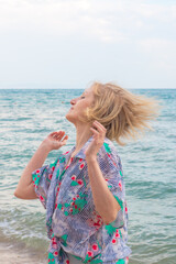 Woman with short blonde hair throwing head back on beach with ocean in background. Carefree summer seaside vacation.