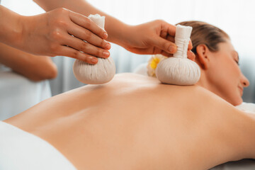 Hot herbal ball spa massage body treatment, masseur gently compresses herb bag on couple customer body. Tranquil and serenity of aromatherapy recreation in day lighting ambient at spa salon. Quiescent