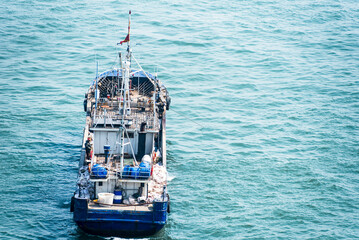 A blue and white fishing boat sails on a calm sea, with gentle waves. The vessel features a deck equipped with fishing gear and a visible wheelhouse.