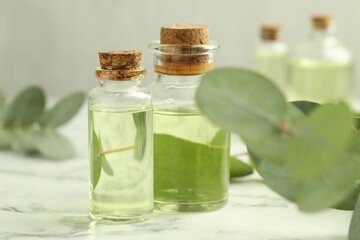 Bottles of essential oil and eucalyptus leaves on white marble table, closeup