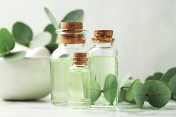 Bottles of essential oil, eucalyptus leaves, mortar and pestle on white marble table, closeup