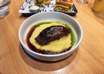 Slow-Cooked Beef Cheek on Creamy Mashed Potatoes with Rich Gravy and Herb Oil.Gourmet Plated Dish: Tender Braised Meat with Potato Puree, Dark Jus, and Pesto.Hearty Restaurant Meal