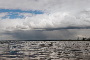 Dramatic clouds loom over a lake shoreline, with distant rainfall visible on the horizon and a hint of blue sky breaking through ample copy space.