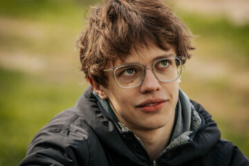 A young man in a black jacket and glasses sits by a wooden table outdoors, appearing thoughtful and...