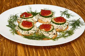 Round canapés with cucumber slices, red caviar and cream filling on dill-decorated plate over wood texture. Concept of elegant holiday appetizer
