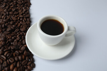Hot coffee in a white coffee cup and many coffee beans scattered around 