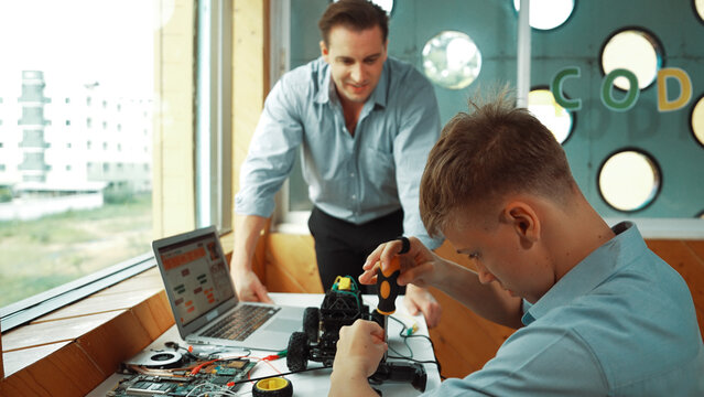 Caucasian teacher help student fixing car model and giving advise for programing system or coding prompt while highschool boy listen and use equipment to repair robot at STEM classroom. Edification.