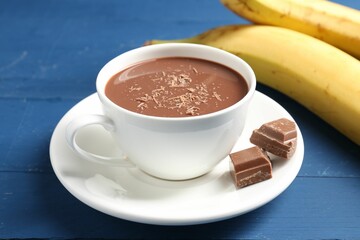 Tasty chocolate milk in cup, pieces and bananas on blue wooden table, closeup