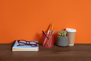 Stylish workplace with stationery, coffee and plant on wooden desk near orange wall. Space for text