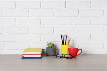 Different stationery, coffee, glasses and floral decor on wooden office table near white brick wall