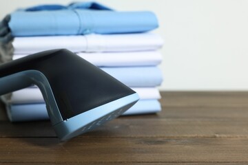 Upright steamer and stack of folded shirts on wooden table near white wall indoors, closeup. Space...