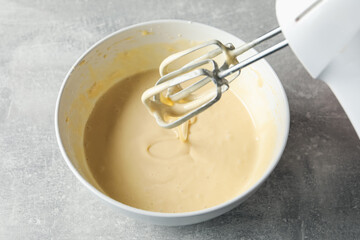 Hand mixer with whisks and bowl of dough on grey textured table, closeup