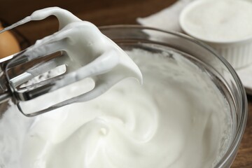 Hand mixer with whisks and bowl of whipped cream on wooden table, closeup