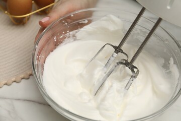 Woman whisking cream with hand mixer at white marble table, closeup