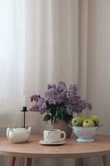 A beautifully serene still life featuring lilacs, vibrant red apples, and a charming teapot on a rustic wooden table.