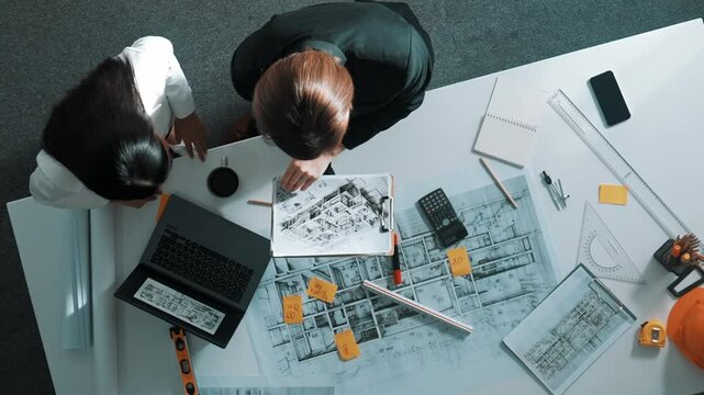Top view of skilled project manager holding project plan and explain house design to architect engineer at meeting table with equipment, ruler, safety helmet and laptop scatter around. Alimentation.
