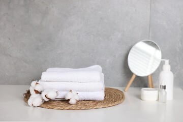 Stack of clean towels, cotton flowers, cosmetic products and mirror on white table indoors