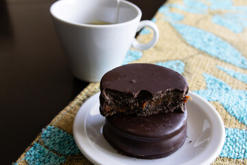 A bitten dark chocolate alfajor on top of a whole alfajor on a white plate next to a cup of tea
