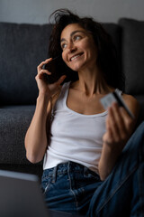 Fototapeta premium Smiling young woman sitting on couch at home, holding credit card and talking on smartphone while making online payment or shopping as a freelancer with a laptop.