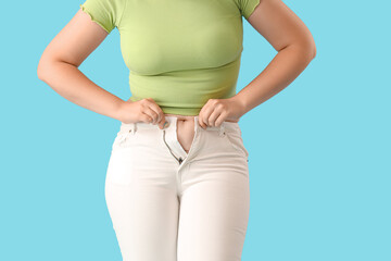 Young woman trying to button tight jeans on blue background, closeup. Weight gain concept